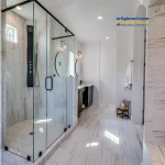 Bagno luminoso con doccia walk-in in vetro, rivestimento effetto marmo e piatto con mosaico; vasca freestanding accanto alla finestra.